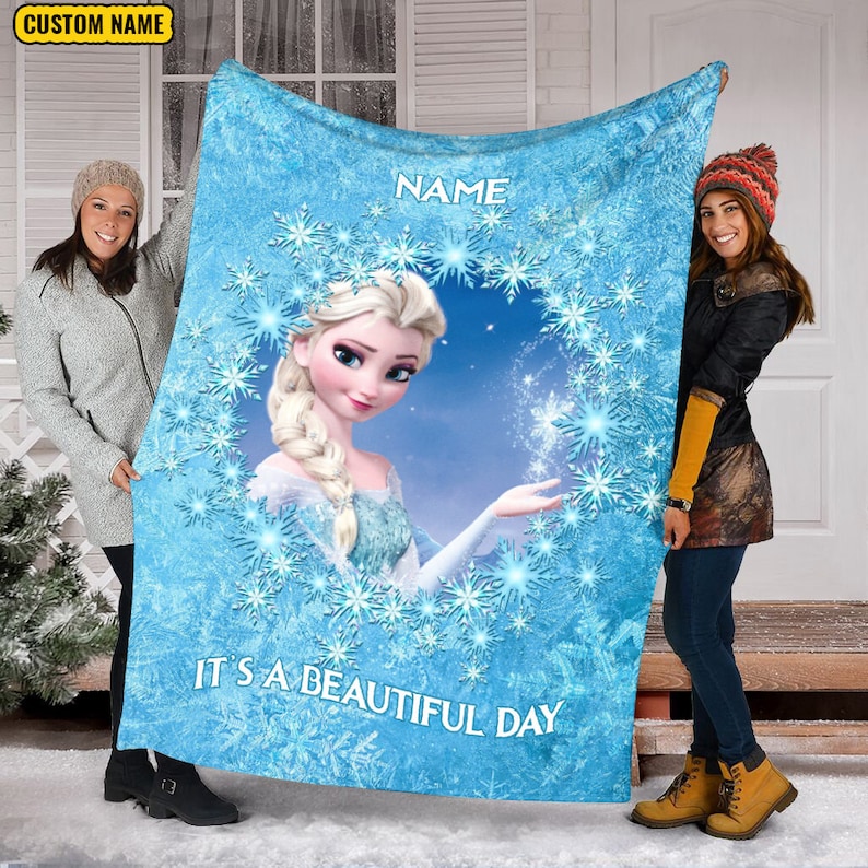 Pu&ograve; includere: Una coperta in pile azzurro con un'illustrazione centrale di un personaggio biondo con una treccia, circondato da una corona di fiocchi di neve blu. Il testo "NAME" appare sopra e "IT'S A BEAUTIFUL DAY" &egrave; stampato sotto.