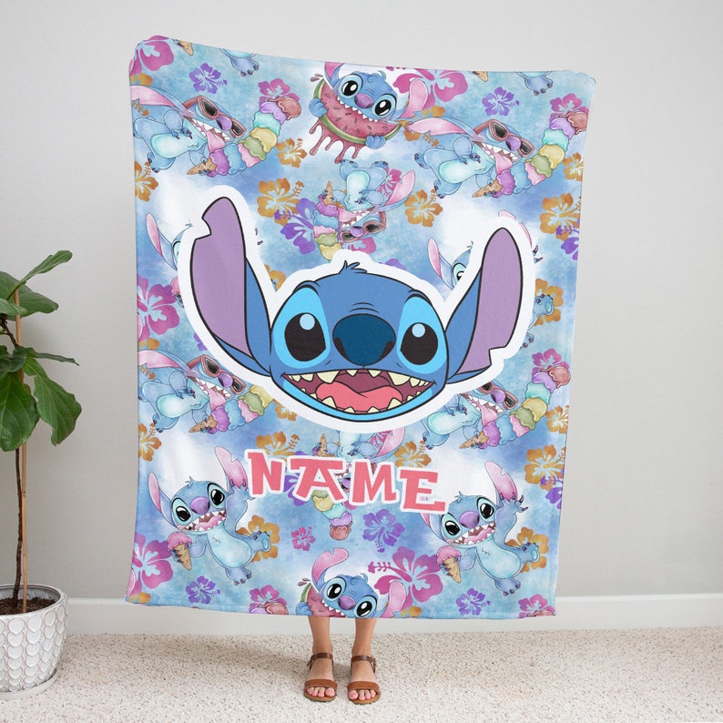 Puede incluir: Una manta de forro polar suave con un patr&oacute;n repetitivo del personaje azul Stitch comiendo helado y sand&iacute;a, intercalado con flores de hibisco rosas y naranjas sobre un fondo azul. Aparece una cara grande de Stitch y el texto NAME.