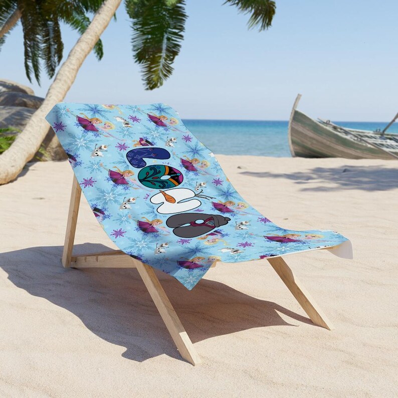 May include: A light blue beach chair with a patterned towel featuring the word "Snow" in colorful letters. The towel has a snowflake and character design. The chair is made of wood and is on a sandy beach.