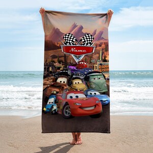 May include: A rectangular beach towel featuring characters from the animated movie "Cars." The design includes Lightning McQueen, Mater, and other characters, with a "Name" banner and checkered flags. The background depicts a desert landscape.