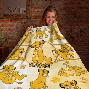 May include: A white fleece blanket with a yellow and brown pattern of various cartoon lions from the movie The Lion King. The blanket has the word "NAME" printed in the center.