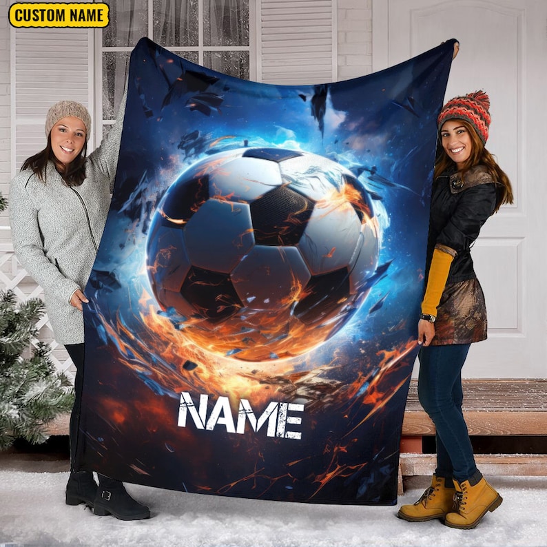 May include: A black fleece blanket with a soccer ball engulfed in flames. The ball is white with black panels. The background is a dark blue with orange and yellow flames. The text "NAME" is printed in white on the blanket.