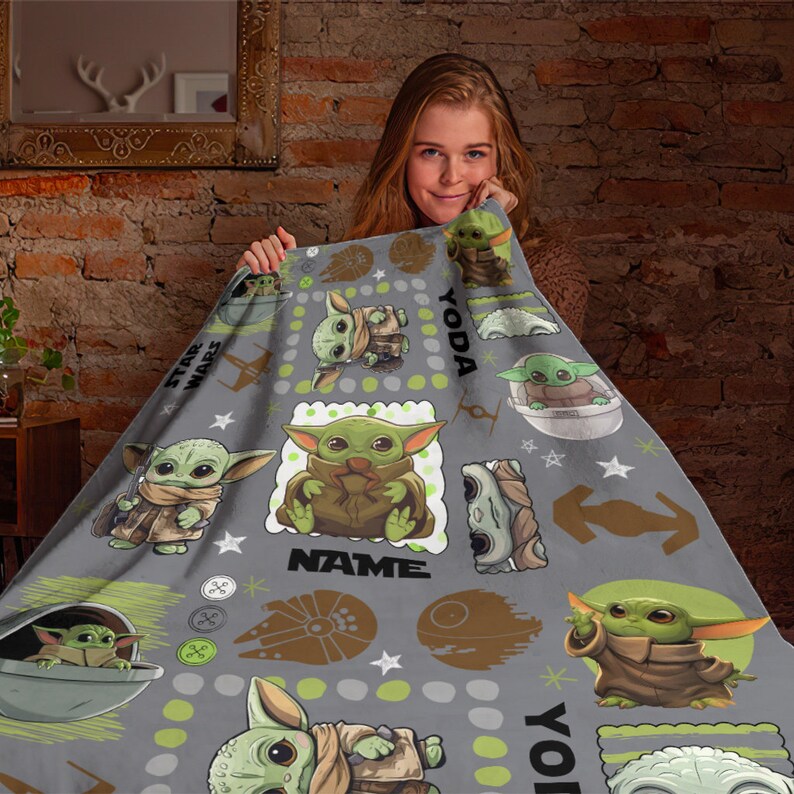 May include: A gray fleece blanket featuring a repeating pattern of the green character Grogu from Star Wars in various poses, including inside a floating pod. The design includes the text YODA and NAME, along with brown icons of spaceships.