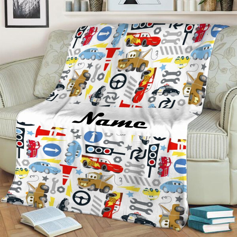 May include: A white fleece blanket featuring a repeating pattern of cartoon race cars, tow trucks, police cars, traffic lights, road signs, wrenches, and lightning bolts. The word Name is printed in a black cursive font across the centre.