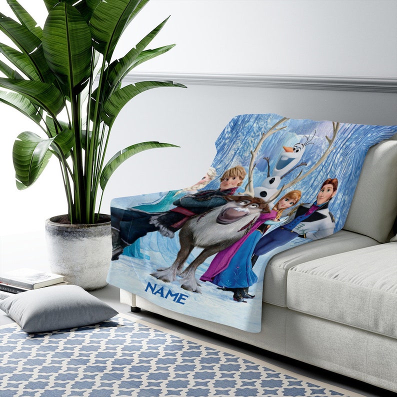 May include: A soft throw blanket featuring a colorful graphic print of characters from the movie Frozen, including Anna, Elsa, Kristoff, Sven, and Olaf against a blue icy background. The word NAME is printed in blue at the bottom of the blanket.