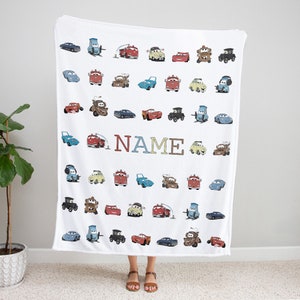 K&ouml;nnte beinhalten: Eine wei&szlig;e Fleecedecke mit einem sich wiederholenden Muster aus verschiedenen Zeichentrick-Autofiguren aus dem Film Cars, die in Reihen angeordnet sind. Das Wort NAME ist in gro&szlig;en, bunten Blockbuchstaben in der Mitte des Designs gedruckt.