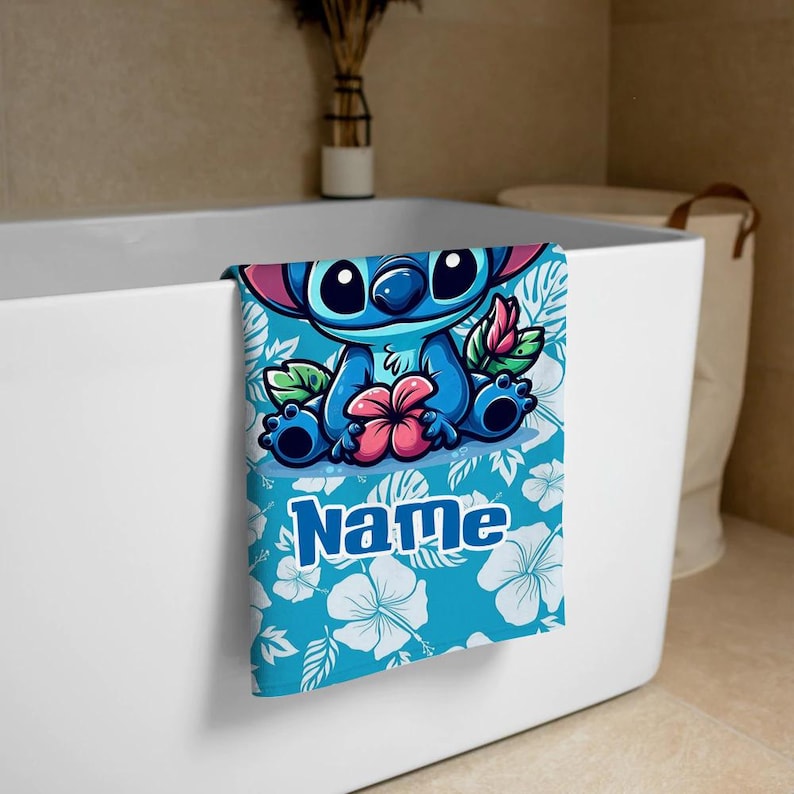 May include: A blue and white towel featuring a cartoon character with large eyes, a pink flower, and the word "Name". The towel has a blue background with white floral patterns and is draped over a white bathtub.