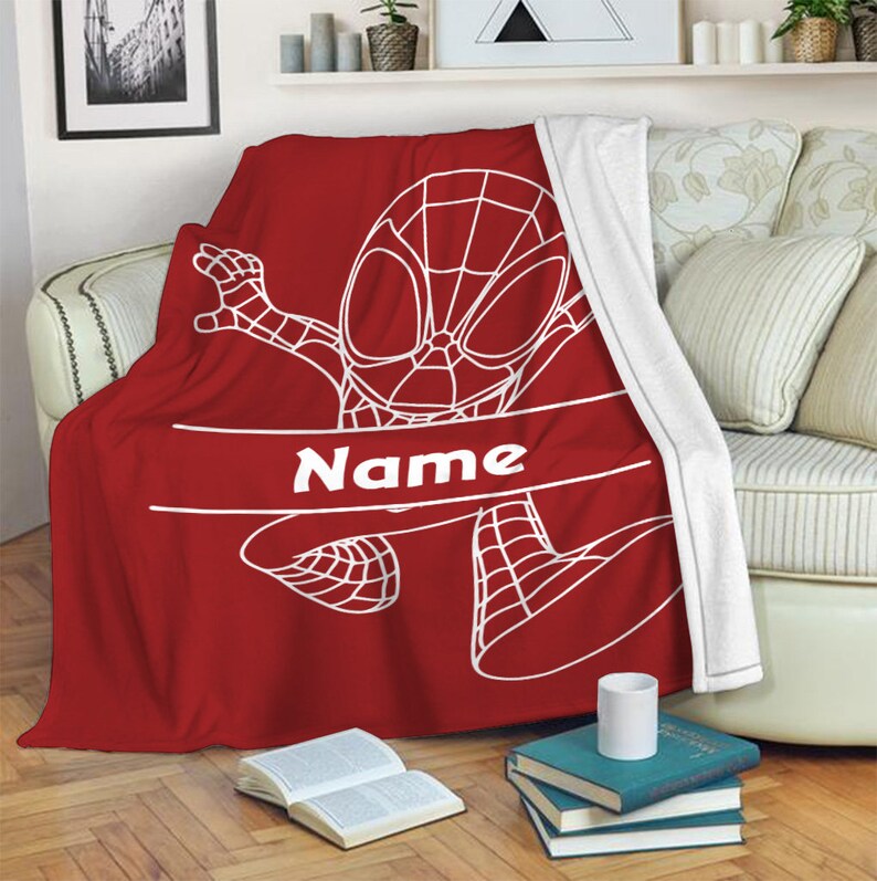 May include: A red fleece blanket featuring a white line-art illustration of a superhero character in a webbed mask and suit. A horizontal white banner across the center contains the word Name in white text. The blanket has a white plush lining.