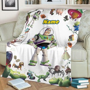 Puede incluir: Una manta de forro polar blanca con un gran gr&aacute;fico central de Buzz Lightyear rodeado de varios personajes de Toy Story, incluyendo a Woody, Rex, Slinky Dog y los Aliens. La palabra Name est&aacute; impresa en amarillo con un borde azul arriba.