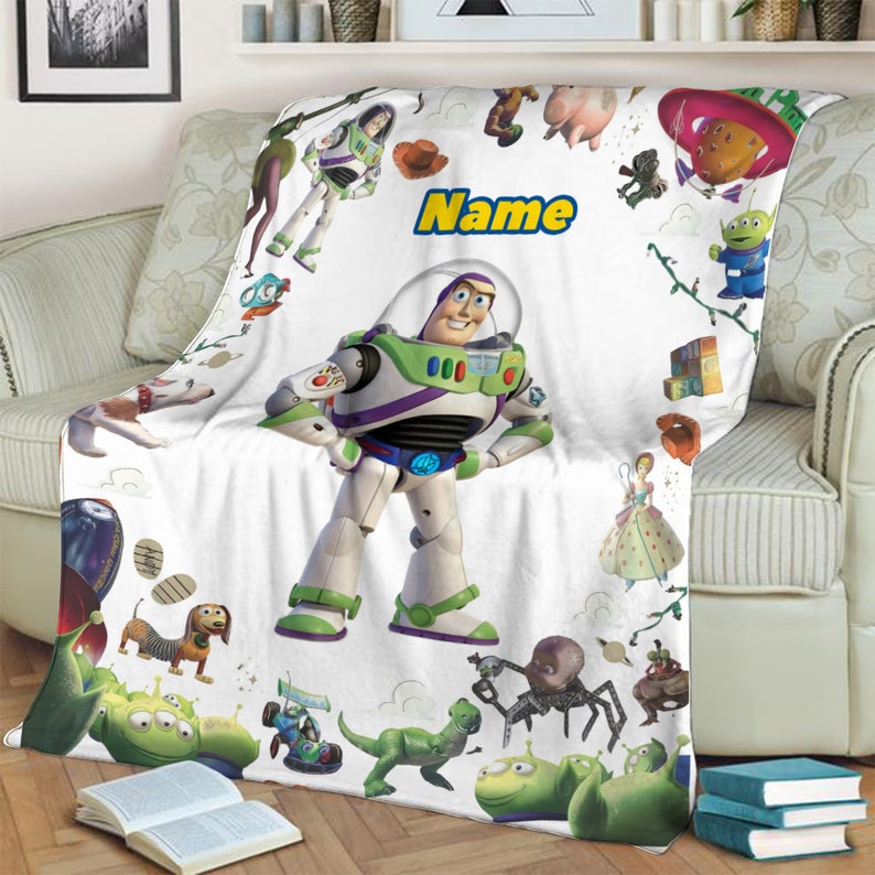 Puede incluir: Una manta de forro polar blanca con un gran gr&aacute;fico central de Buzz Lightyear rodeado de varios personajes de Toy Story, incluyendo a Woody, Rex, Slinky Dog y los Aliens. La palabra Name est&aacute; impresa en amarillo con un borde azul arriba.