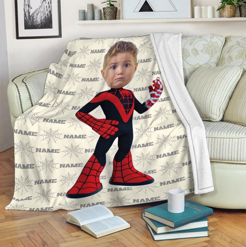 May include: A cream-coloured throw blanket featuring a repeating pattern of grey spiderwebs and the word NAME in grey text. Centered on the blanket is a cartoon illustration of a child with a superhero body in a red and black suit.