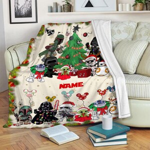 Puede incluir: Una manta de forro polar blanca con un dise&ntilde;o navide&ntilde;o con personajes de Star Wars. Los personajes est&aacute;n vestidos con atuendos festivos y se re&uacute;nen alrededor de un &aacute;rbol de Navidad. La manta tiene un nombre personalizado, "NAME", impreso en ella.