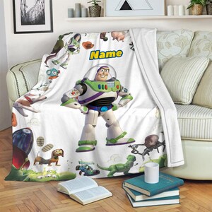 Puede incluir: Una manta de forro polar blanca con un gran gr&aacute;fico central del personaje Buzz Lightyear, rodeado de varios personajes de Toy Story, incluyendo un perro salchicha, un dinosaurio verde y un cohete, con la palabra Name impresa en amarillo.
