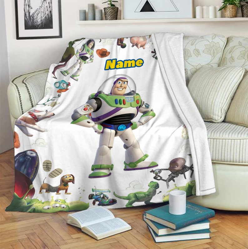 Puede incluir: Una manta de forro polar blanca con un gran gr&aacute;fico central del personaje Buzz Lightyear, rodeado de varios personajes de Toy Story, incluyendo un perro salchicha, un dinosaurio verde y un cohete, con la palabra Name impresa en amarillo.