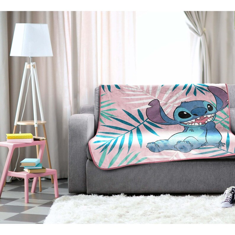 K&ouml;nnte beinhalten: Eine rosa Decke mit einem Cartoon-Bild von Stitch aus Lilo & Stitch. Die Decke hat ein tropisches Bl&auml;ttermuster in T&uuml;rkis und Wei&szlig;. Die Decke ist &uuml;ber ein graues Sofa drapiert.