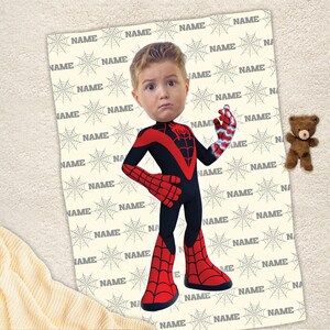May include: A rectangular blanket featuring a repeating pattern of grey spiderwebs and the word NAME in grey text. Centered on the blanket is a cartoon-style illustration of a child with a superhero body in a red and black spider-themed suit.