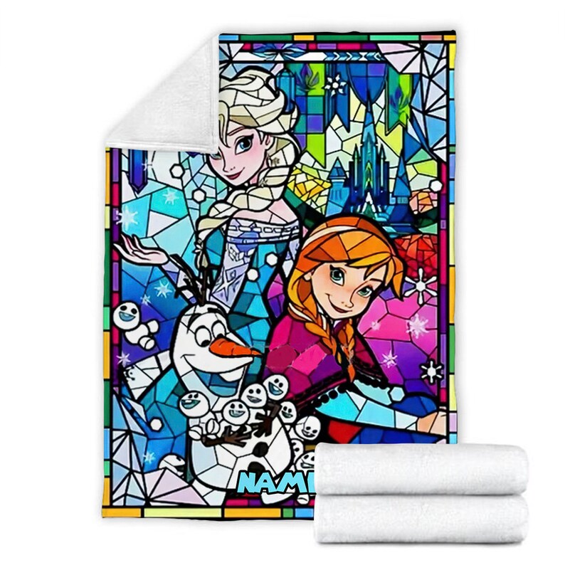 Pode incluir: Uma manta retangular de tecido polar com uma estampa colorida estilo vitral de Elsa, Anna e Olaf de Frozen. O design inclui um castelo de gelo azul, flocos de neve e o nome parcial NAME em letras azuis na parte inferior. A manta &eacute; branca.