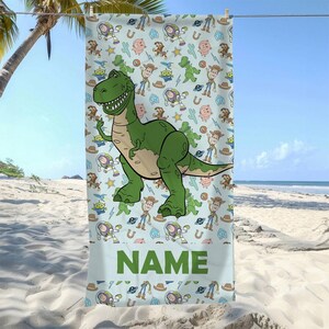 May include: A light blue beach towel featuring a repeating pattern of cartoon characters including a large green dinosaur, space rangers, cowboys, and pigs, with the word NAME printed in large green capital letters at the bottom section.