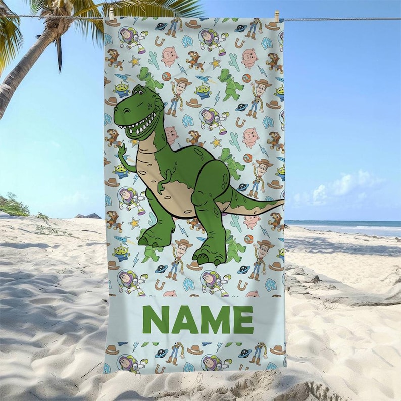 May include: A light blue beach towel featuring a repeating pattern of cartoon characters including a large green dinosaur, space rangers, cowboys, and pigs, with the word NAME printed in large green capital letters at the bottom section.