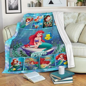 K&ouml;nnte beinhalten: Eine weiche Fleecedecke mit einer Collage aus Szenen von Arielle, die Meerjungfrau, mit Arielle, Fabius und Sebastian. Das Design zeigt ein gro&szlig;es zentrales Bild von Arielle auf einem Felsen mit dem Text NAME, umgeben von kleineren Unterwasserszenen.