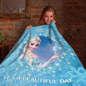 Pu&ograve; includere: Una coperta in pile azzurro con un motivo circolare centrale raffigurante un personaggio biondo con una treccia, circondato da un bordo di fiocchi di neve. Il testo "NAME" appare in alto e "IT'S A BEAUTIFUL DAY" &egrave; stampato in basso.