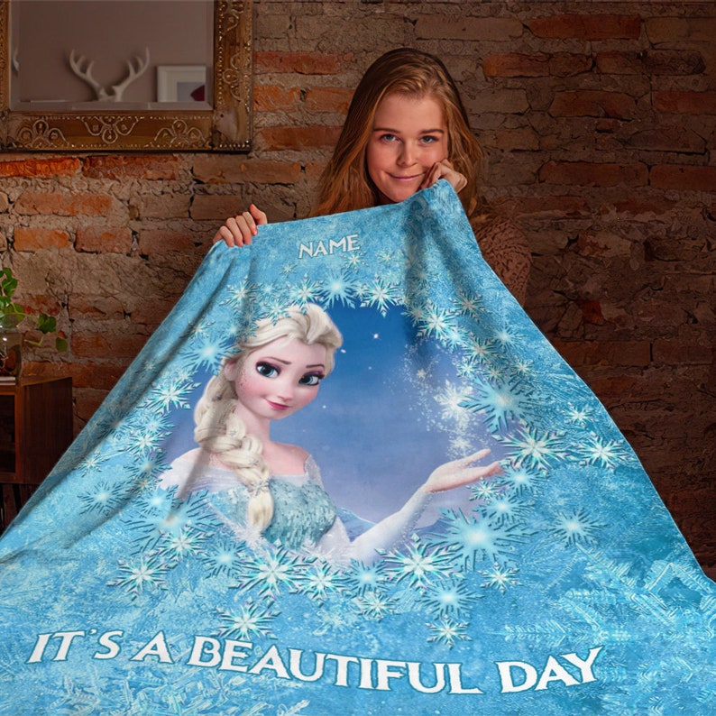 Pu&ograve; includere: Una coperta in pile azzurro con un motivo circolare centrale raffigurante un personaggio biondo con una treccia, circondato da un bordo di fiocchi di neve. Il testo "NAME" appare in alto e "IT'S A BEAUTIFUL DAY" &egrave; stampato in basso.