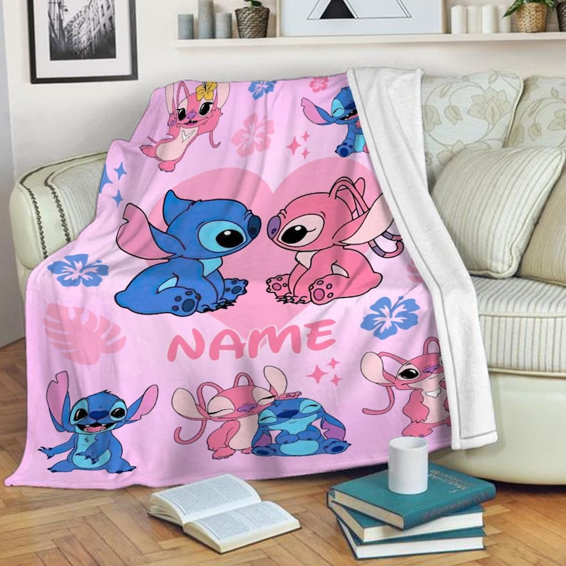 May include: Pink throw blanket featuring cartoon characters Stitch and Angel, with a heart design and the word "NAME". The blanket has a white backing and is draped over a sofa.