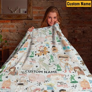 K&ouml;nnte beinhalten: Eine wei&szlig;e Fleecedecke mit einem wiederkehrenden Muster aus Zeichentrickfiguren, darunter ein Cowboy, ein Weltraum-Ranger, ein Dinosaurier, ein Schwein und Plastiksoldaten. Der Text "CUSTOM NAME" und "ANDY'S ROOM KEEP OUT" ist aufgedruckt.