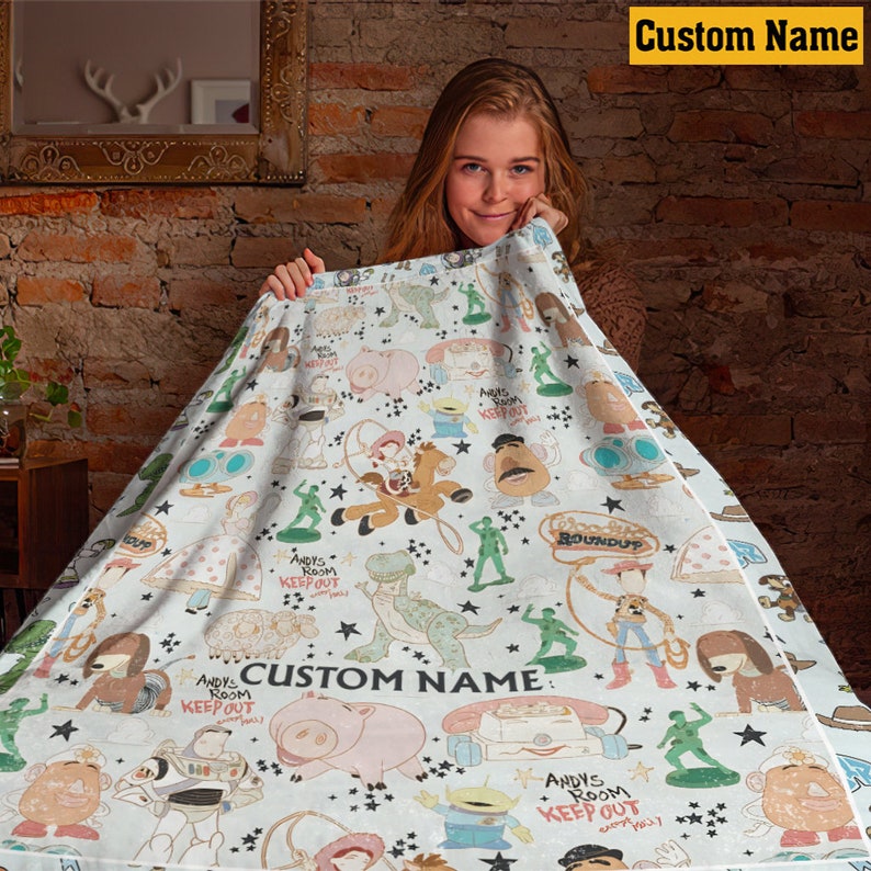 K&ouml;nnte beinhalten: Eine wei&szlig;e Fleecedecke mit einem wiederkehrenden Muster aus Zeichentrickfiguren, darunter ein Cowboy, ein Weltraum-Ranger, ein Dinosaurier, ein Schwein und Plastiksoldaten. Der Text "CUSTOM NAME" und "ANDY'S ROOM KEEP OUT" ist aufgedruckt.