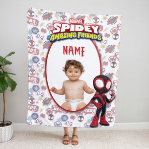 May include: A white fleece blanket featuring a Marvel Spidey and His Amazing Friends pattern with red and blue spider icons and text. The center displays a large oval frame containing a photograph of a baby in a diaper, with the word NAME above.