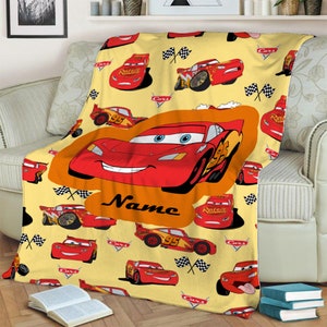 Może przedstawiać: Ż&oacute;łty koc polarowy z powtarzającym się wzorem czerwonych samochod&oacute;w wyścigowych Lightning McQueen z filmu Auta, flagami w szachownicę i logo Cars. Duży centralny pomarańczowy owal podkreśla dużą grafikę samochodu ze słowem Name poniżej.