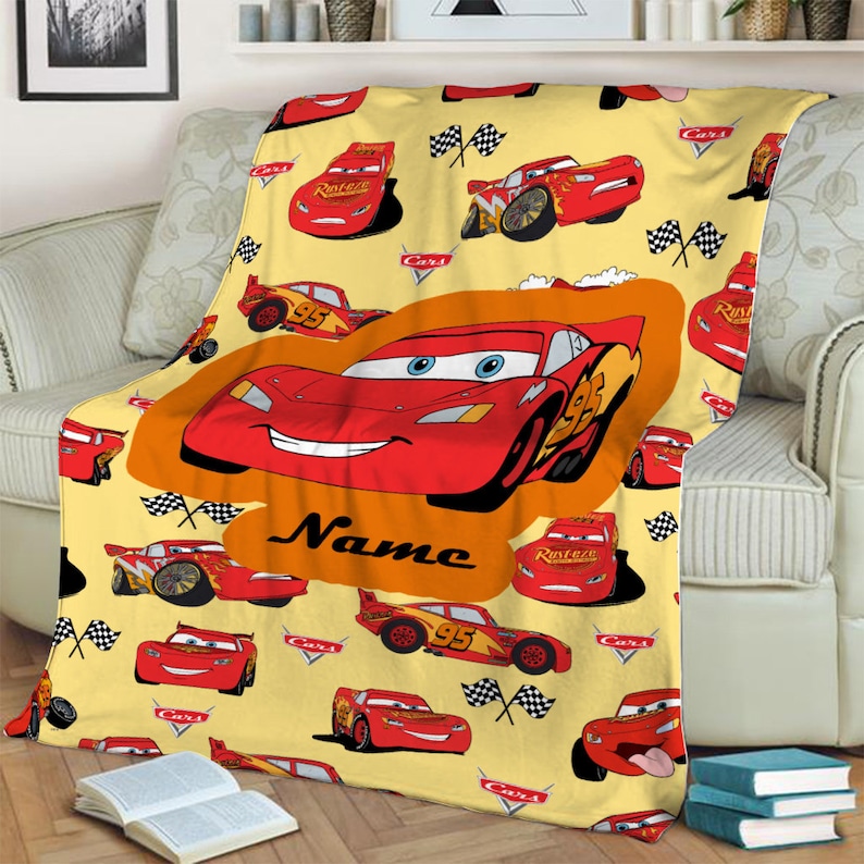 Może przedstawiać: Ż&oacute;łty koc polarowy z powtarzającym się wzorem czerwonych samochod&oacute;w wyścigowych Lightning McQueen z filmu Auta, flagami w szachownicę i logo Cars. Duży centralny pomarańczowy owal podkreśla dużą grafikę samochodu ze słowem Name poniżej.