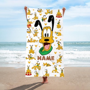 May include: White beach towel featuring a repeating pattern of Pluto cartoon characters in various poses, with a large Pluto face and the word "NAME" in brown. The towel is held up against a beach and ocean backdrop.