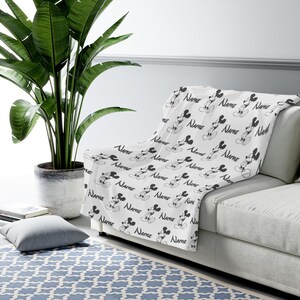 May include: A white throw blanket featuring a repeating black and white pattern of Mickey Mouse illustrations and the word Name printed in a script font. The blanket is draped over a light-colored sofa, showcasing the consistent graphic design.