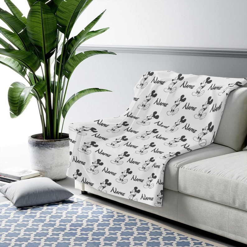 May include: A white throw blanket featuring a repeating black and white pattern of Mickey Mouse illustrations and the word Name printed in a script font. The blanket is draped over a light-colored sofa, showcasing the consistent graphic design.