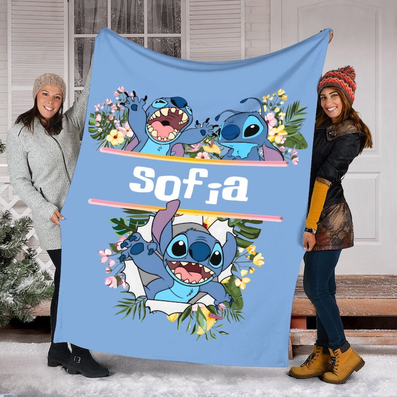 May include: A light blue fleece blanket featuring three illustrations of the blue character Stitch surrounded by tropical flowers and green leaves. The name Sofia is printed in white block letters in the center between two horizontal pink lines.