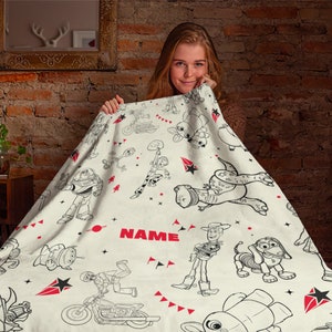 Peut inclure: Une couverture en polaire cr&egrave;me avec un motif en lignes noires repr&eacute;sentant divers personnages anim&eacute;s, dont un ranger de l'espace, un cow-boy, un dinosaure et un chien. Le design inclut des &eacute;toiles rouges et le mot NAME en texte rouge.