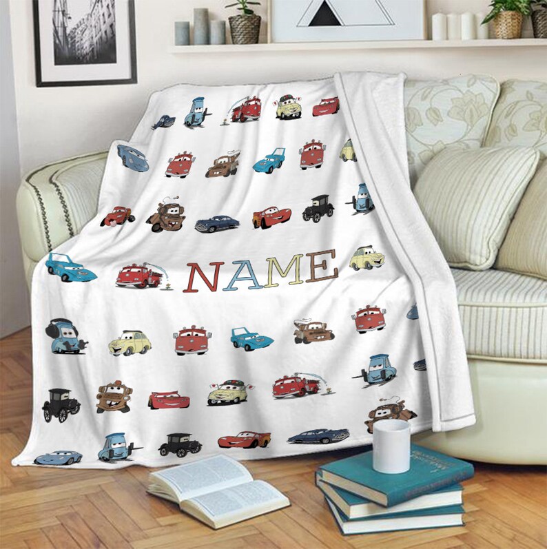 K&ouml;nnte beinhalten: Eine wei&szlig;e Fleecedecke mit einem sich wiederholenden Muster aus verschiedenen bunten Zeichentrickautos und Lastwagen aus dem Film Cars, wobei das Wort NAME in gro&szlig;en, mehrfarbigen Blockbuchstaben in der Mitte des Stoffes gedruckt ist.