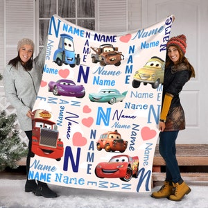 May include: A white fleece blanket featuring a repeating pattern of characters from the movie Cars, including Lightning McQueen, Mater, and others. The design incorporates the word Name in blue text, the letter N, and pink hearts throughout.