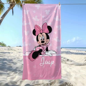 Puede incluir: Una toalla de playa rectangular en tonos rosa con un patr&oacute;n repetitivo de siluetas de Minnie Mouse y estrellas blancas. El centro muestra un gr&aacute;fico de Minnie Mouse con un vestido de lunares y un lazo rosa, con el nombre Julie en letra cursiva blanca.