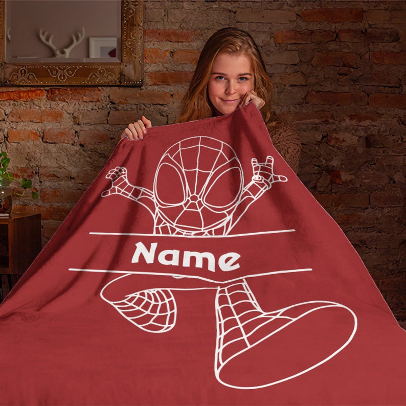 May include: A deep red fleece blanket featuring a white line art illustration of a superhero character in a webbed suit. The design includes a horizontal banner across the center with the word Name printed in bold white capital letters.