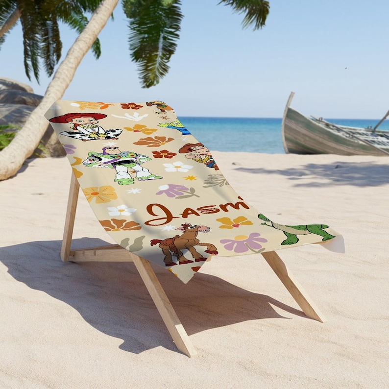 May include: A beige beach towel featuring a repeating pattern of Toy Story characters including Buzz Lightyear, Jessie, Woody, Bullseye, and Rex, interspersed with colorful floral motifs and the name Jasmine printed in a brown cursive script.