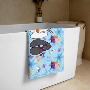 May include: A light blue bath towel featuring cartoon characters and snowflakes. The towel has images of Olaf, Elsa, and Anna from the movie Frozen. The towel is folded over the edge of a white bathtub.