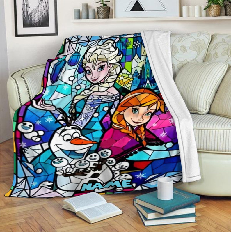 Pode incluir: Uma manta de pel&uacute;cia com uma estampa colorida estilo vitral de Elsa, Anna e Olaf de Frozen. O design inclui padr&otilde;es geom&eacute;tricos em azul, roxo e rosa com a palavra NAME impressa em azul claro na parte central inferior.
