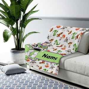 May include: A white fleece throw blanket featuring a repeating pattern of Toy Story characters including Buzz Lightyear, Woody, and Rex. A large green horizontal band across the center displays the word Name in bold black script lettering.
