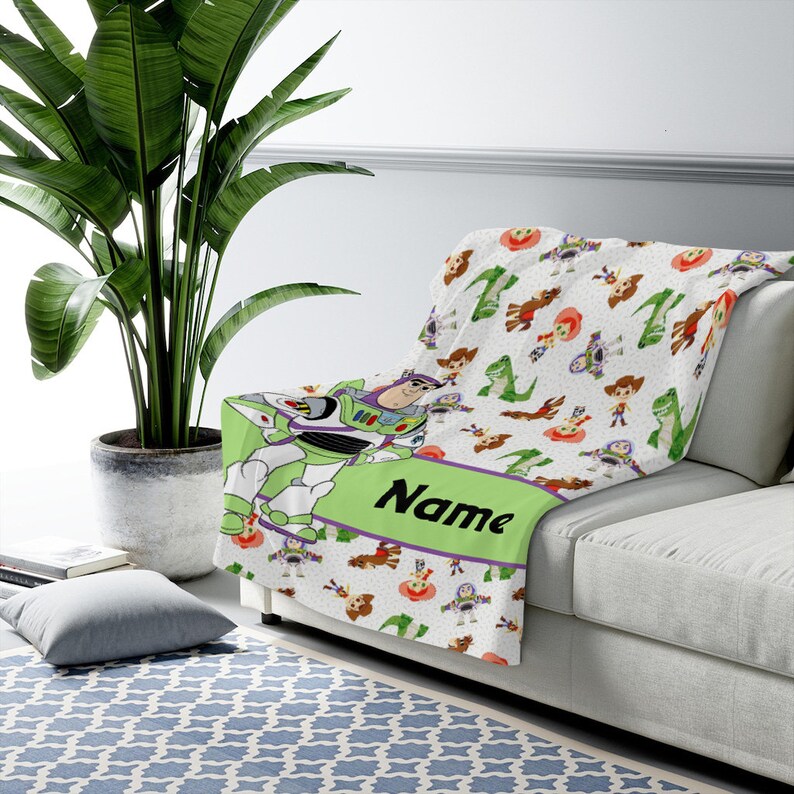 May include: A white fleece throw blanket featuring a repeating pattern of Toy Story characters including Buzz Lightyear, Woody, and Rex. A large green horizontal band across the center displays the word Name in bold black script lettering.