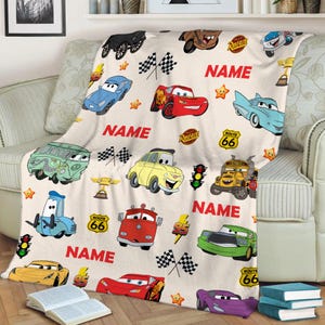 May include: A white fleece blanket with a Cars theme. The blanket features various Cars characters, including Lightning McQueen, Mater, and the other cars from the movie. The blanket also features the text "NAME" and "ROUTE 66".