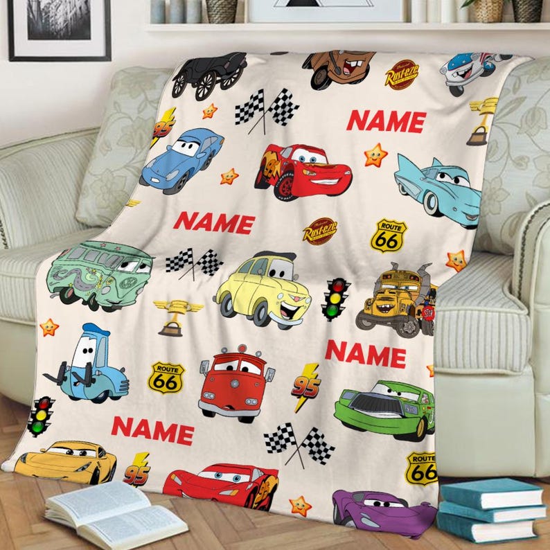 May include: A white fleece blanket with a Cars theme. The blanket features various Cars characters, including Lightning McQueen, Mater, and the other cars from the movie. The blanket also features the text "NAME" and "ROUTE 66".