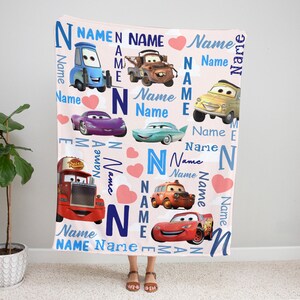 May include: A soft fleece blanket featuring a light pink background with various characters from the Cars movie franchise, including Lightning McQueen, Mater, and others. The design includes scattered pink hearts and the repeated word Name in blue.