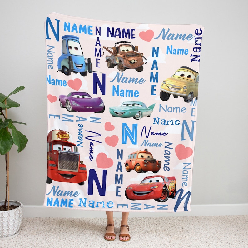 May include: A soft fleece blanket featuring a light pink background with various characters from the Cars movie franchise, including Lightning McQueen, Mater, and others. The design includes scattered pink hearts and the repeated word Name in blue.
